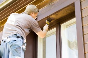 A man with a stands on a ladder applying brown paint to a home's exterior.