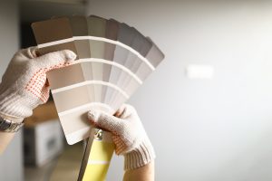 Gloved hands hold up a set of color swatches with earth tones against an interior wall.