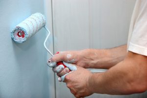 A man uses a roller to apply blue paint to an interior wall.